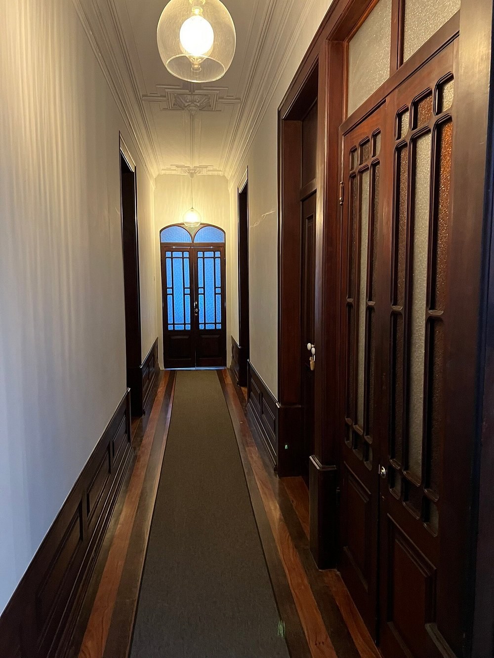 O corredor original da casa, com portas altas de madeira escura, um candeeiro de vidro suspenso e a porta envidraçada ao fundo